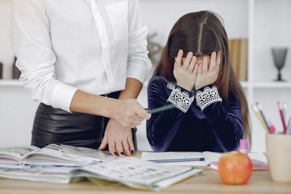 A young girl overwhelmed by homework with a teacher assisting her indoors.