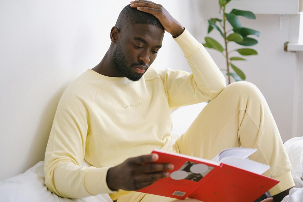 A man in a yellow sweatshirt sits thoughtfully reading a book in a bright indoor setting.